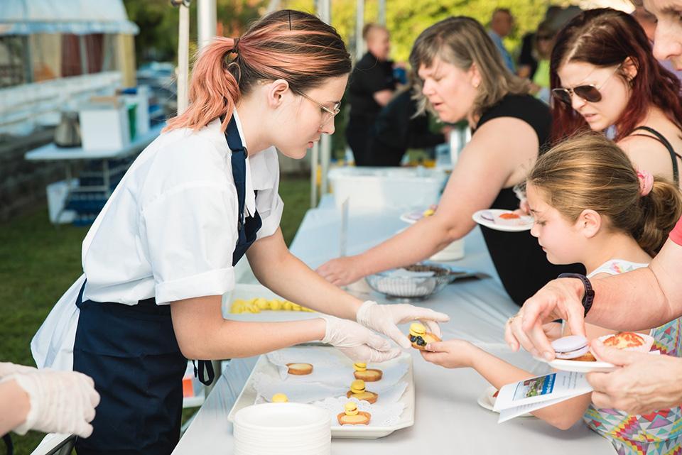 A student wearing an apron and gloves serves a food sample to a young girl attending an event.